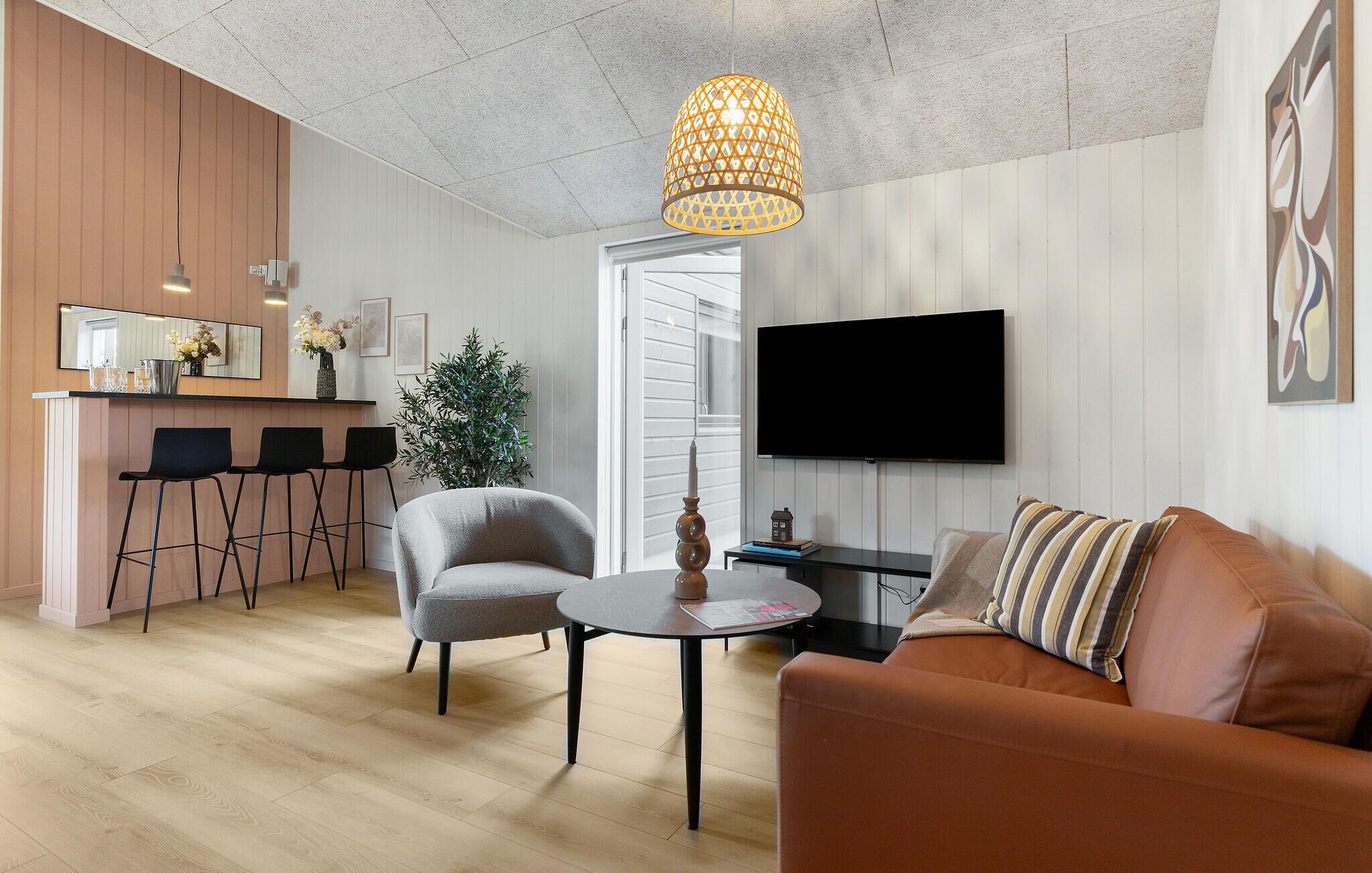 Ett modernt vardagsrum med en grå fåtölj, ett runt soffbord, en brun soffa, en tv och en köksdel med en bardisk och barstolar.