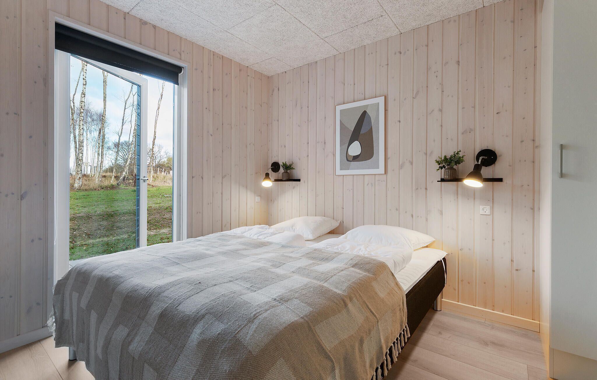 A bedroom with a large window, wooden walls, a bed with white bedding, and two wall-mounted lamps.
