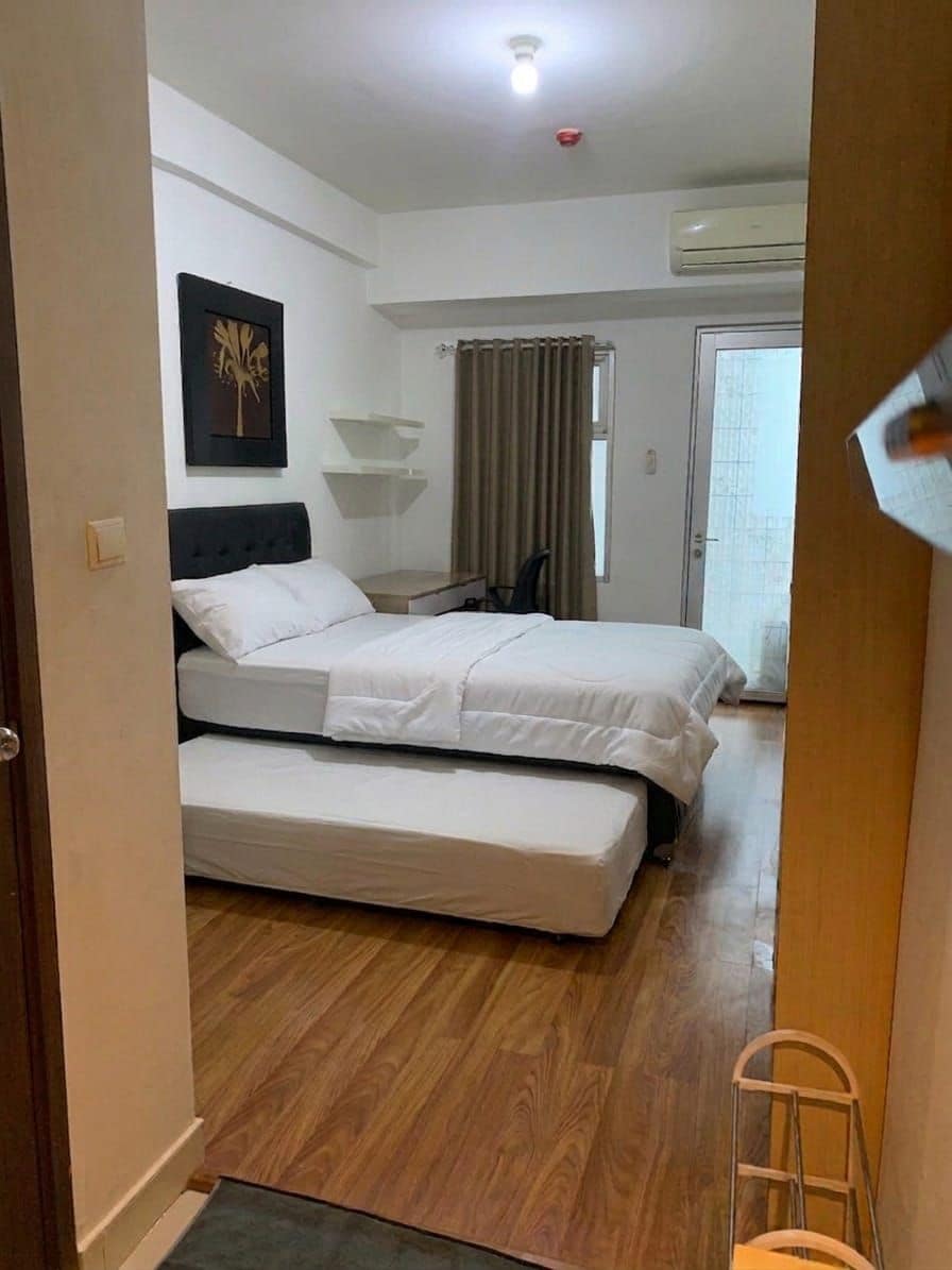 A bedroom with a bed, a bedside table, a chair, a window with curtains, and an air conditioning unit.