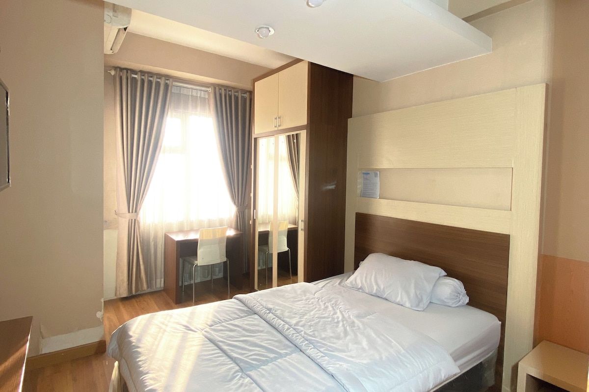 A modern bedroom with a large bed, wooden flooring, a desk with chairs, and a window with curtains.