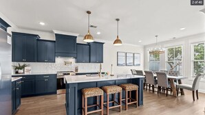 A modern kitchen with dark blue cabinets, a central island with stools, pendant lighting, and a dining area with a large window. - Spacious NEW Luxury 5 Bedroom / 6 Bath Custom Home with Pool and Elevator (North Myrtle Beach)