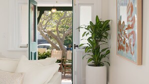 Una stanza con un divano bianco, una pianta in vaso di grandi dimensioni e una porta che dà su uno spazio esterno con un albero e auto parcheggiate.