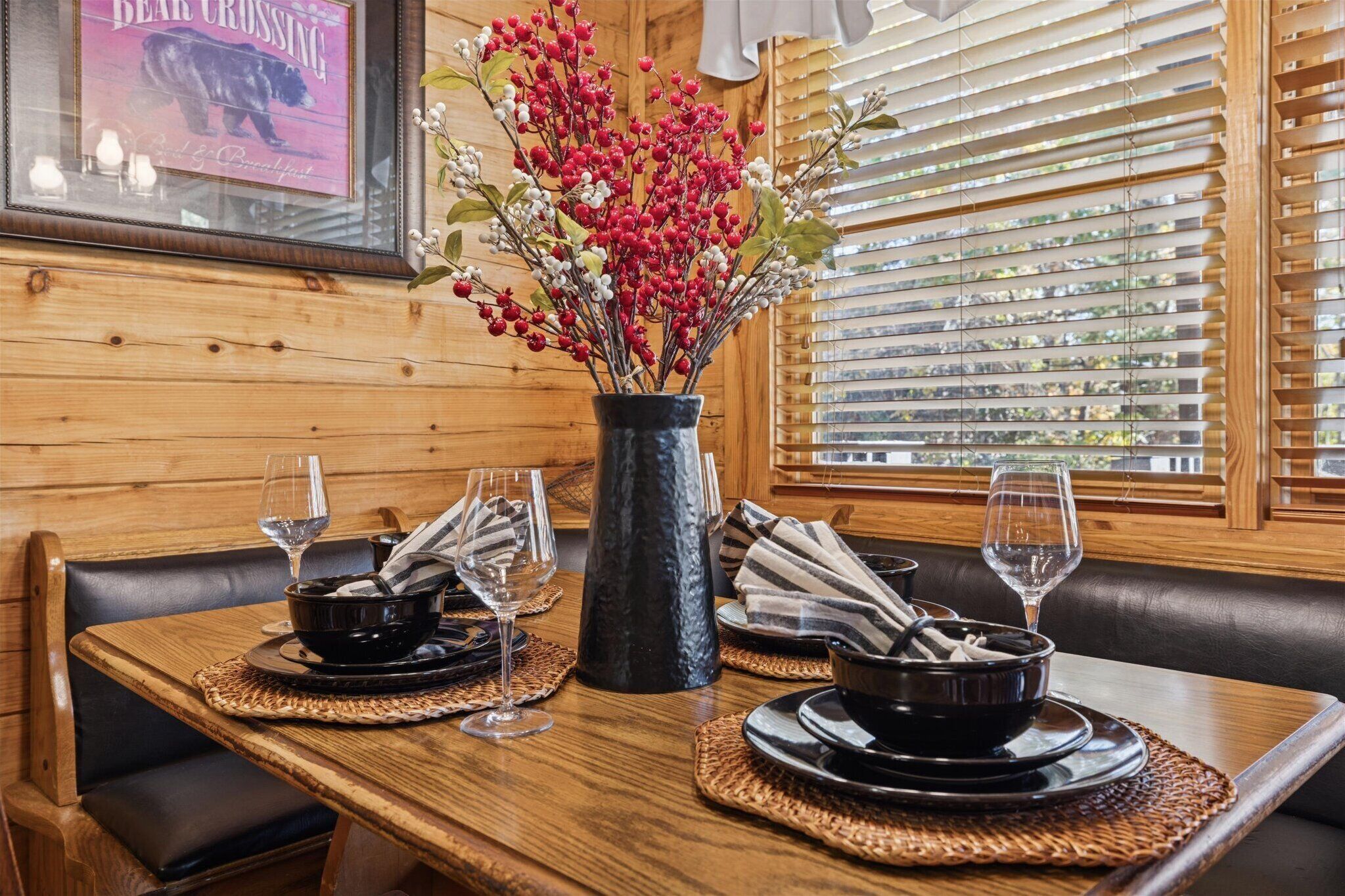 A wooden dining table set with black bowls, wine glasses, and a vase with red berries.