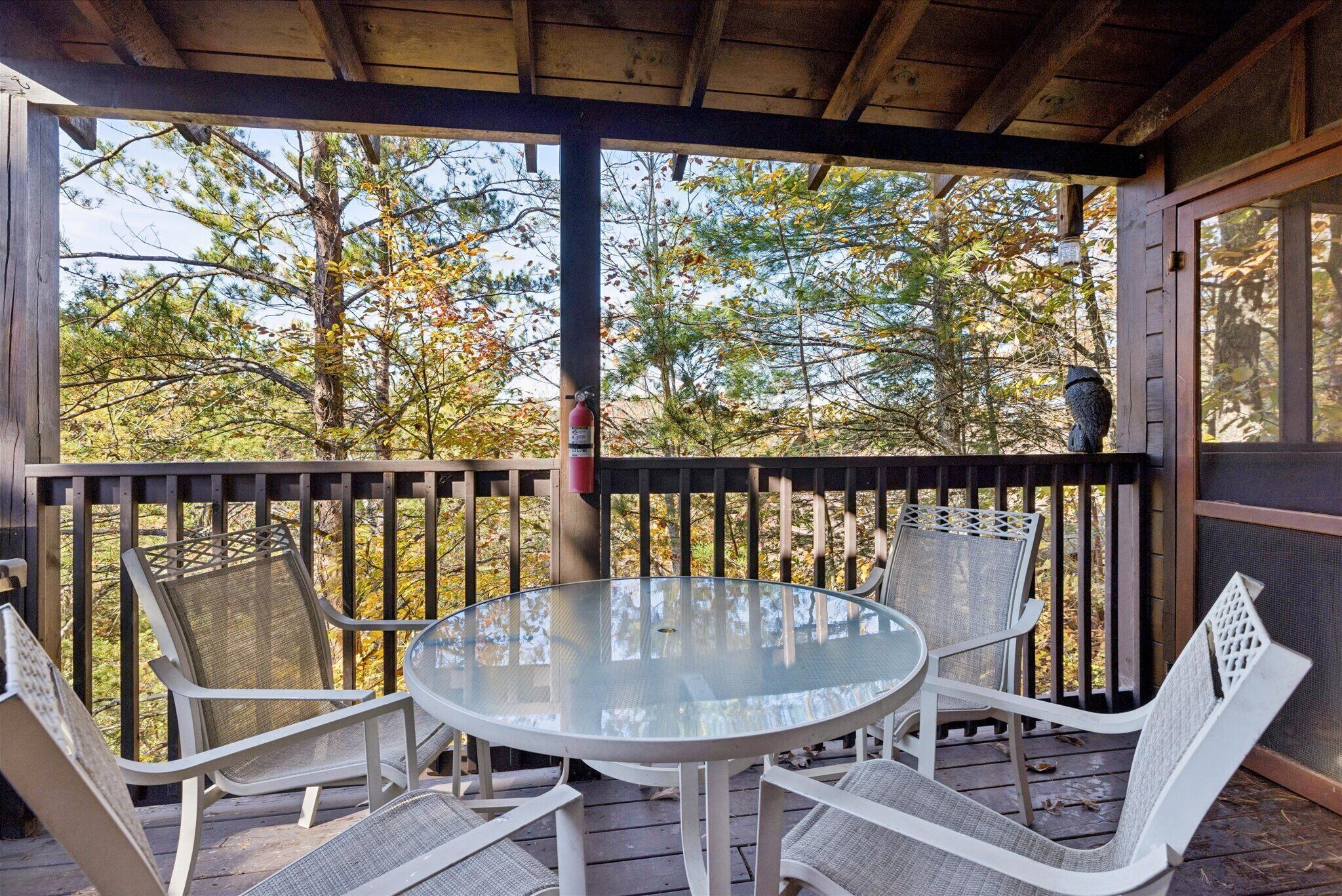 Un espace extérieur aménagé, abrité et doté d’une table ronde en verre et de chaises en osier blanc, entouré d’une terrasse en bois et offrant une vue sur les arbres.
