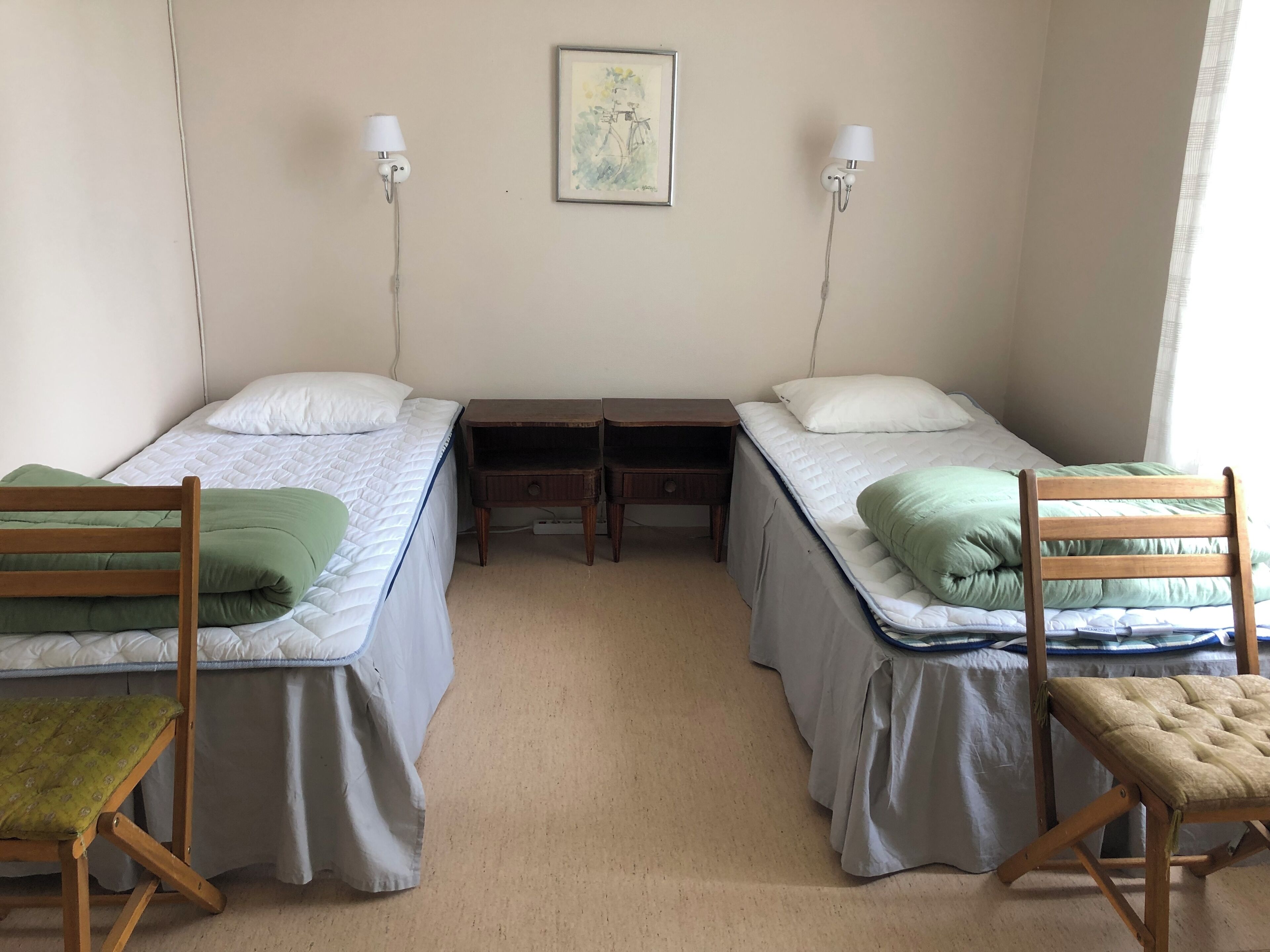 Une chambre avec deux lits simples, chacun équipé d’un oreiller et d’une couverture verte. On y trouve deux tables de chevet avec des lampes, une petite table et un tableau encadré accroché au mur.