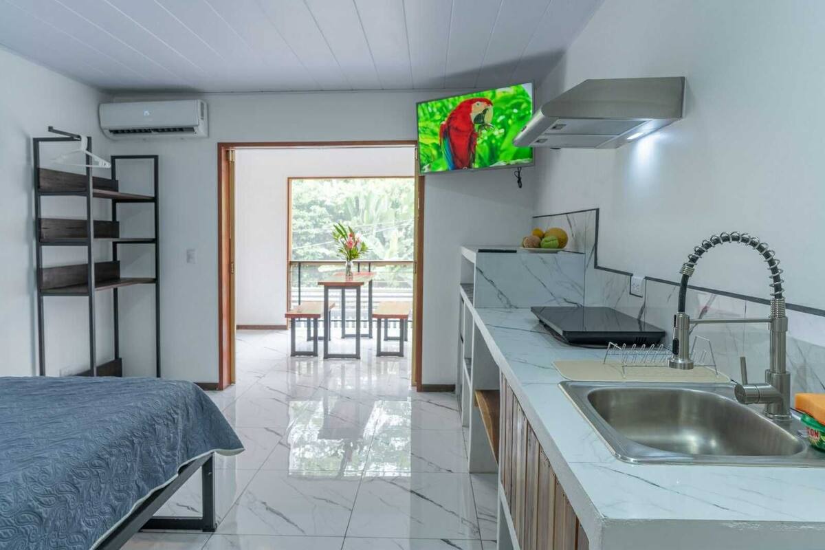 Habitación de hotel compacta con cama, kitchenette, comedor con mesa y sillas, y un ventanal con vistas a zonas verdes.