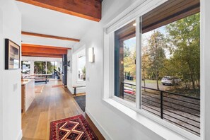 Un hall d’entrée spacieux avec des poutres en bois, une grande fenêtre donnant sur des arbres et une voiture, et un tapis à motifs sur le sol.