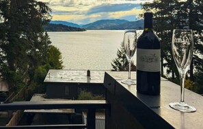 A bottle of wine and two glasses on a balcony overlooking a lake with mountains in the distance.