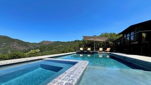 Une piscine aux eaux cristallines, bordée de carreaux, entourée de chaises longues et d’une terrasse en bois, offrant une vue imprenable sur un paysage montagneux.
