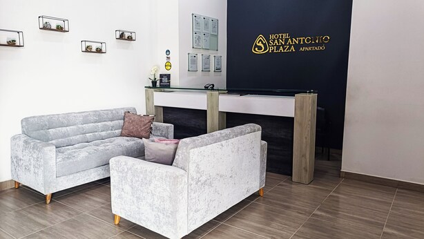 A reception area with a reception desk, two grey sofas, a potted plant, and a wall with a sign for Hotel San Antonio Plaza Apartado. - HOTEL SAN ANTONIO PLAZA APARTADO (Apartadó)
