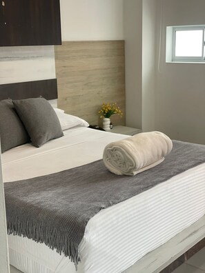 A bed with a grey blanket, a rolled towel, and a vase with yellow flowers on a bedside table.