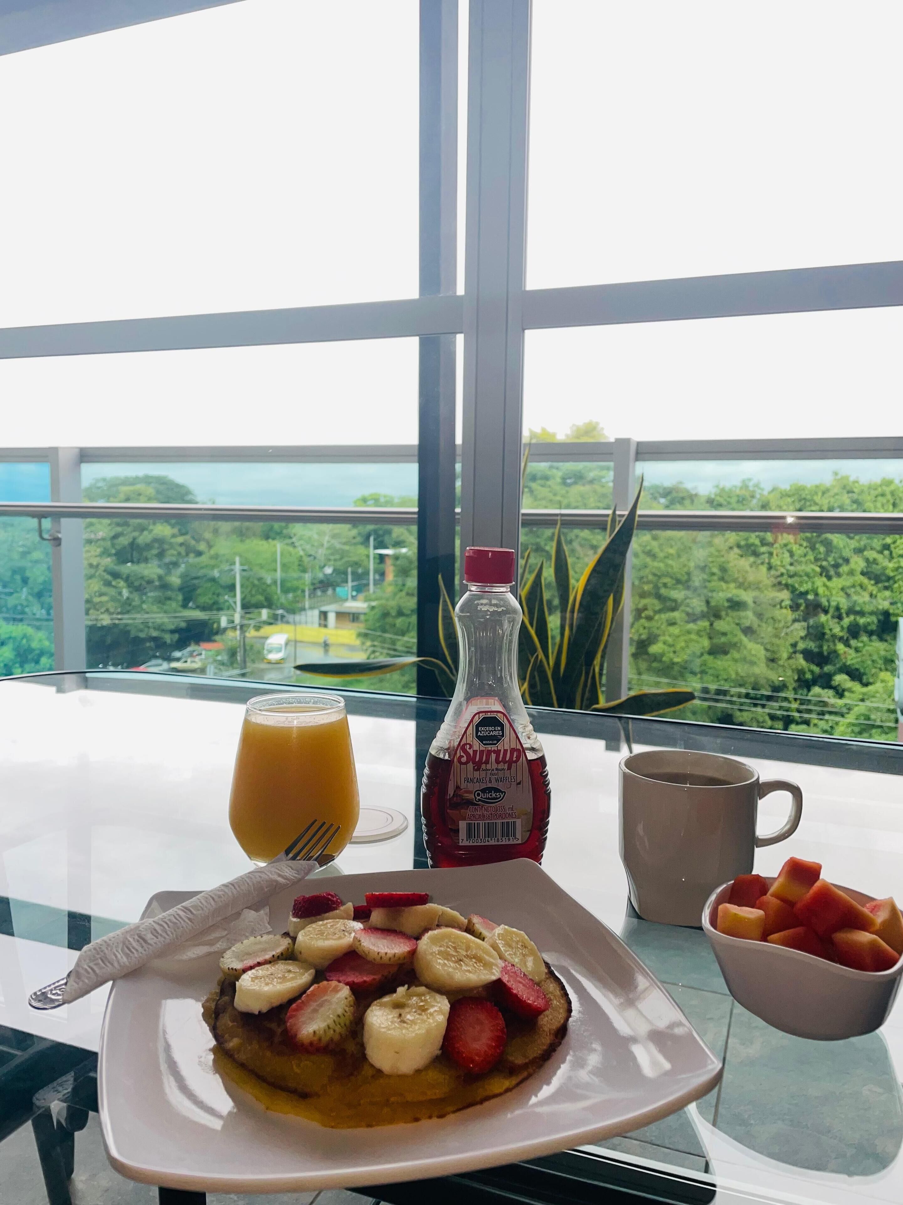 Una mesa de desayuno con tortitas cubiertas de fruta, una botella de jarabe, un vaso de zumo de naranja, una taza de café y un bol de fruta.