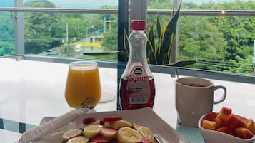 Una mesa de desayuno con tortitas cubiertas de fruta, una botella de jarabe, un vaso de zumo de naranja, una taza de café y un bol de fruta.