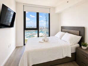 Ein modernes Hotelzimmer mit einem großen Bett, einem an der Wand montierten Fernseher und Blick auf eine Stadtlandschaft durch ein großes Fenster.
