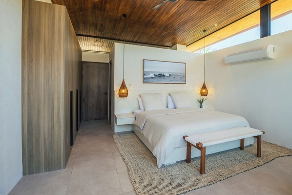 A modern bedroom with a wooden ceiling, a large bed with a white mattress, a bench, and a wall-mounted air conditioner.
