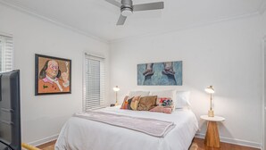 Um quarto com cama, ventoinha no teto, televisão, quadro, abajur e janela com persianas.