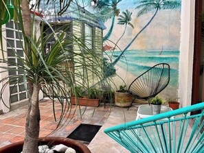 Eine tropisch gestaltete Terrasse mit einer Hängematte, Topfpflanzen und einer Wandmalerei von einer Strandansicht.
