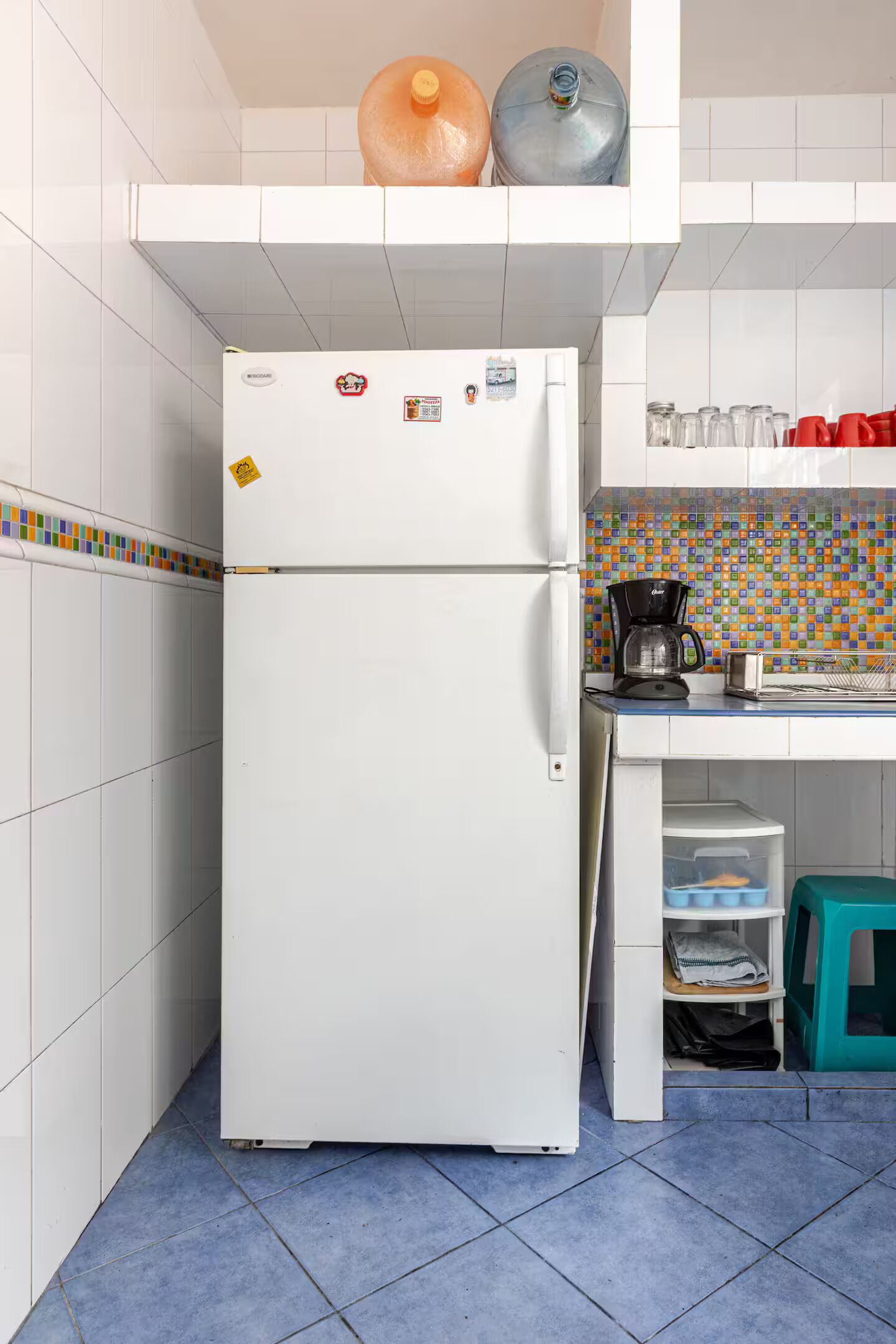 Eine kleine Küche mit einem weißen Kühlschrank, einer Kaffeemaschine und einer farbenfrohen Spritzschutzwand.