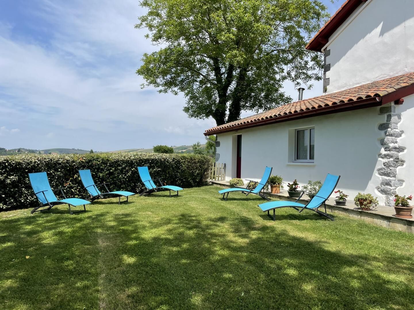 Un bâtiment blanc avec un toit en tuiles et un mur en pierre. Des fauteuils bleus sont disposés en demi-cercle sur une pelouse verdoyante.