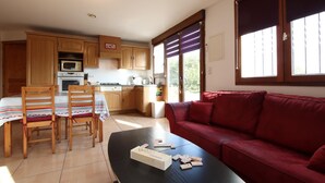 Cocina compacta con armarios de madera, una mesa de comedor con sillas, un sofá rojo y una ventana con persianas.