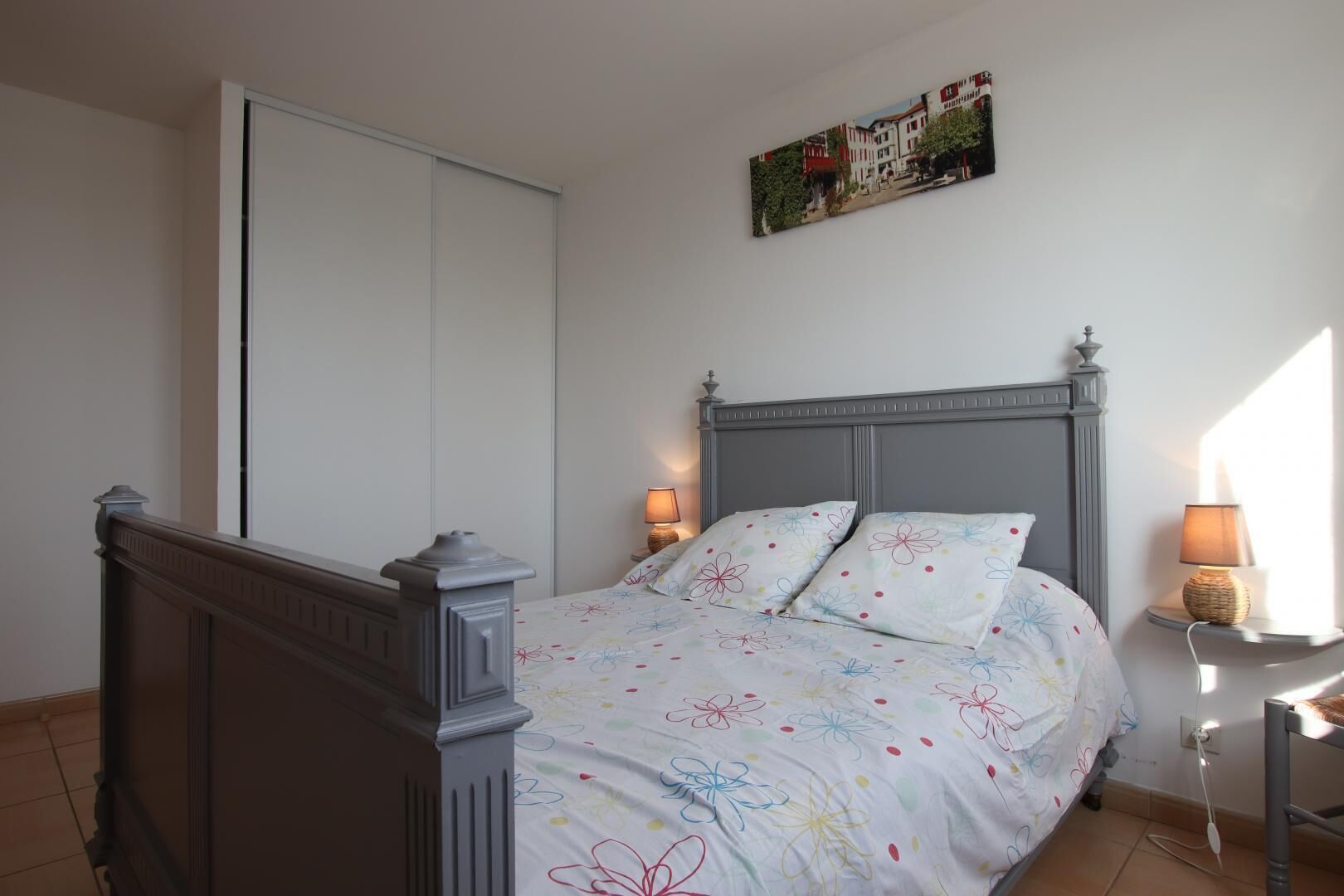 A bedroom with a bed, two bedside lamps, a wall-mounted painting, and a closed door.