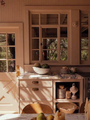 A kitchen with wooden cabinets, a marble countertop, and a window with a view of trees. - A home by designers for design lovers. (Round Top)