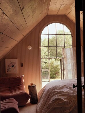 A cozy attic bedroom with a large arched window, wooden walls, a bed, a chair, and a small table. - A home by designers for design lovers. (Round Top)