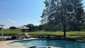 Una piscina con un jacuzzi, rodeada por un deck de piedra y un césped con un árbol y un pabellón blanco.