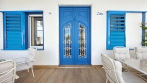 Una habitación con puerta azul, paredes blancas y suelo de madera. Hay dos sillas blancas y una pequeña mesa.