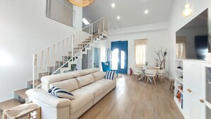 Amplia sala de estar con sofá beige, escalera con baranda de cuerda, mesa de comedor con sillas y área de cocina con armarios.