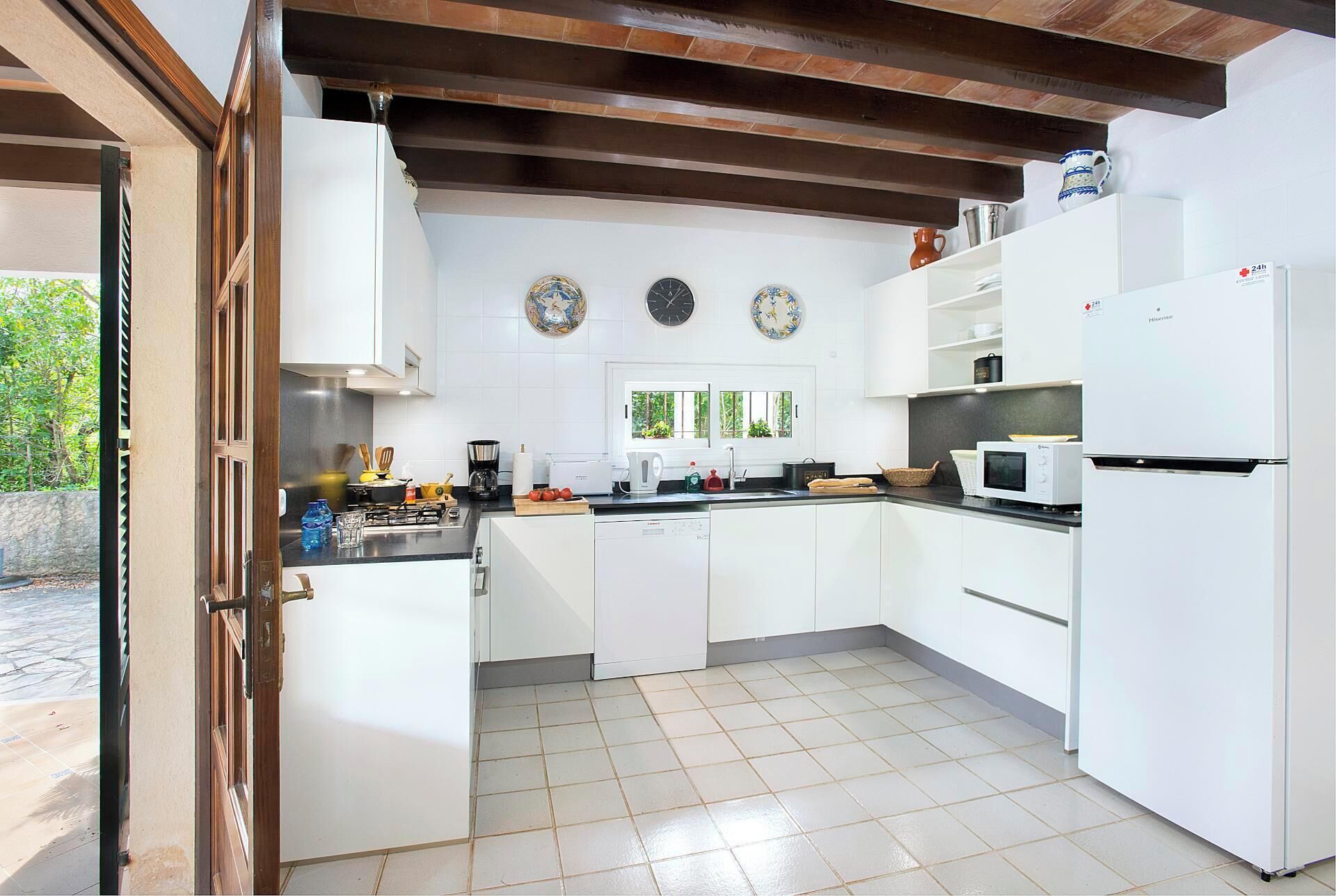 Una cocina moderna con armarios blancos, un microondas empotrado y un refrigerador. Hay dos relojes en la pared, una ventana con vistas a la vegetación y una puerta que da al exterior.