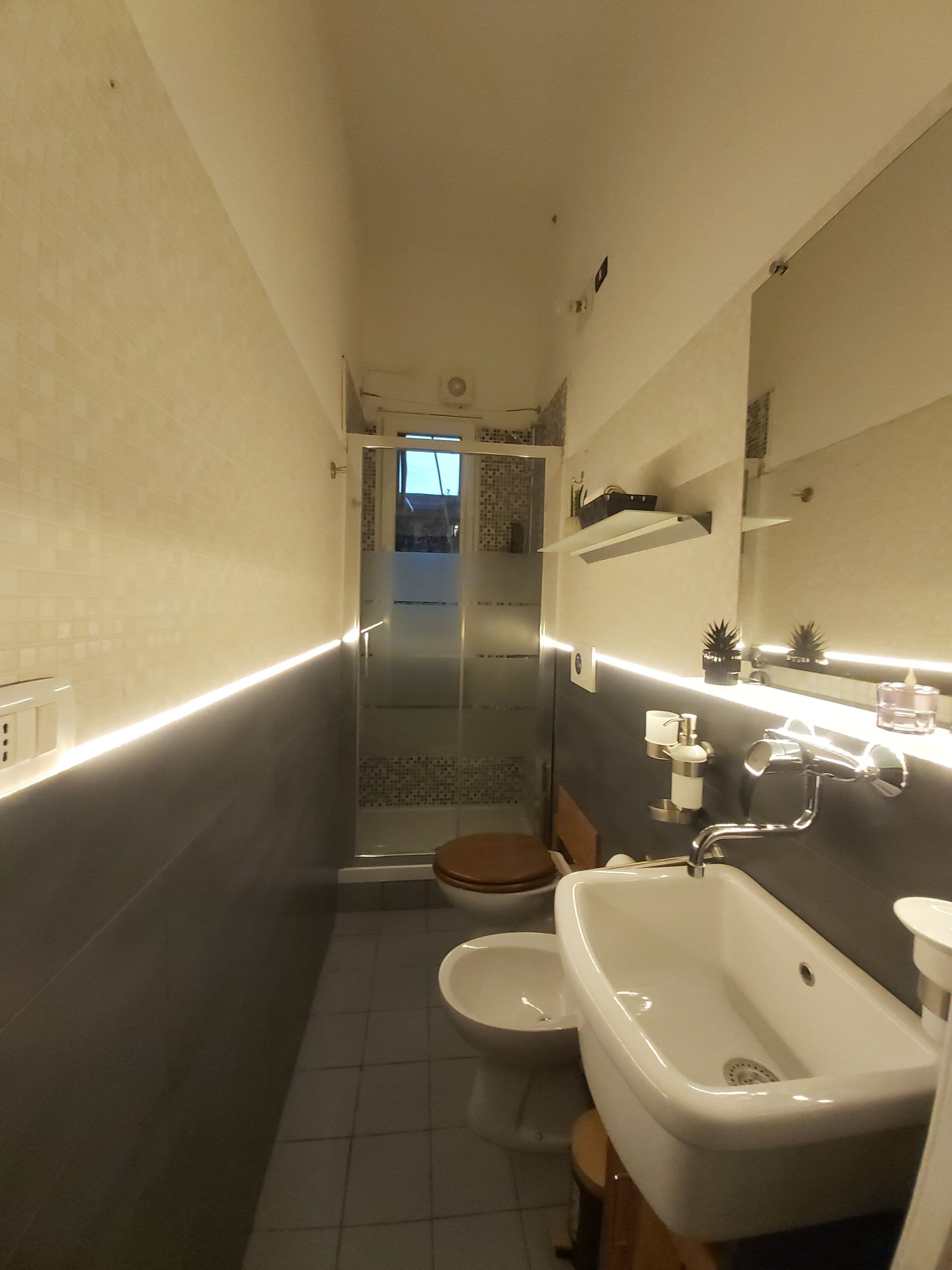 A narrow bathroom with a toilet, bidet, and sink. There is a shower area visible in the background.