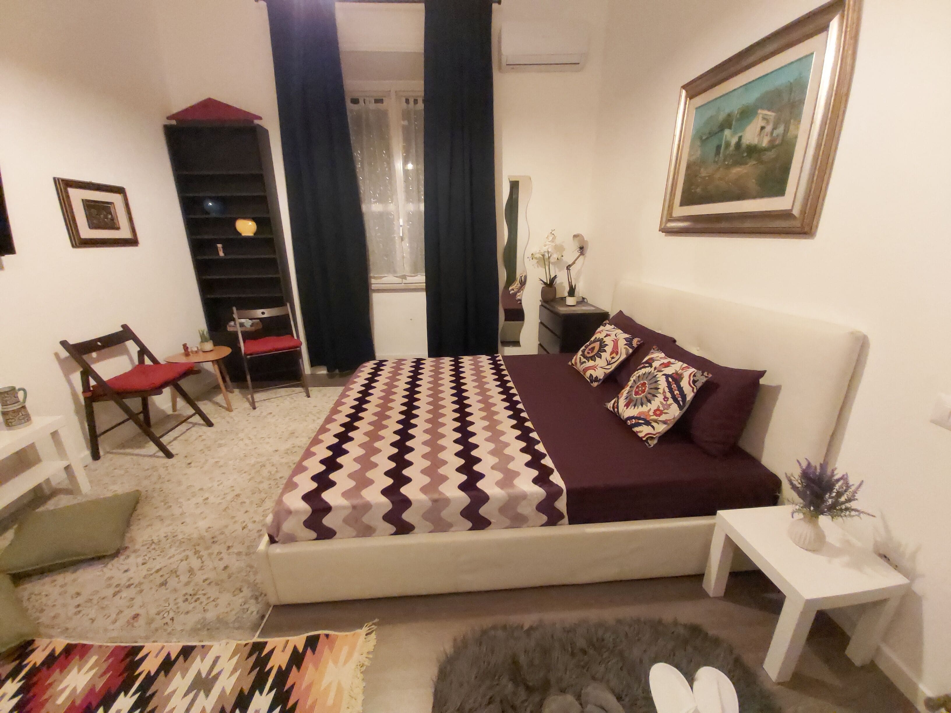 A bedroom with a bed, a chair, a small table, a shelf, and a window with curtains.