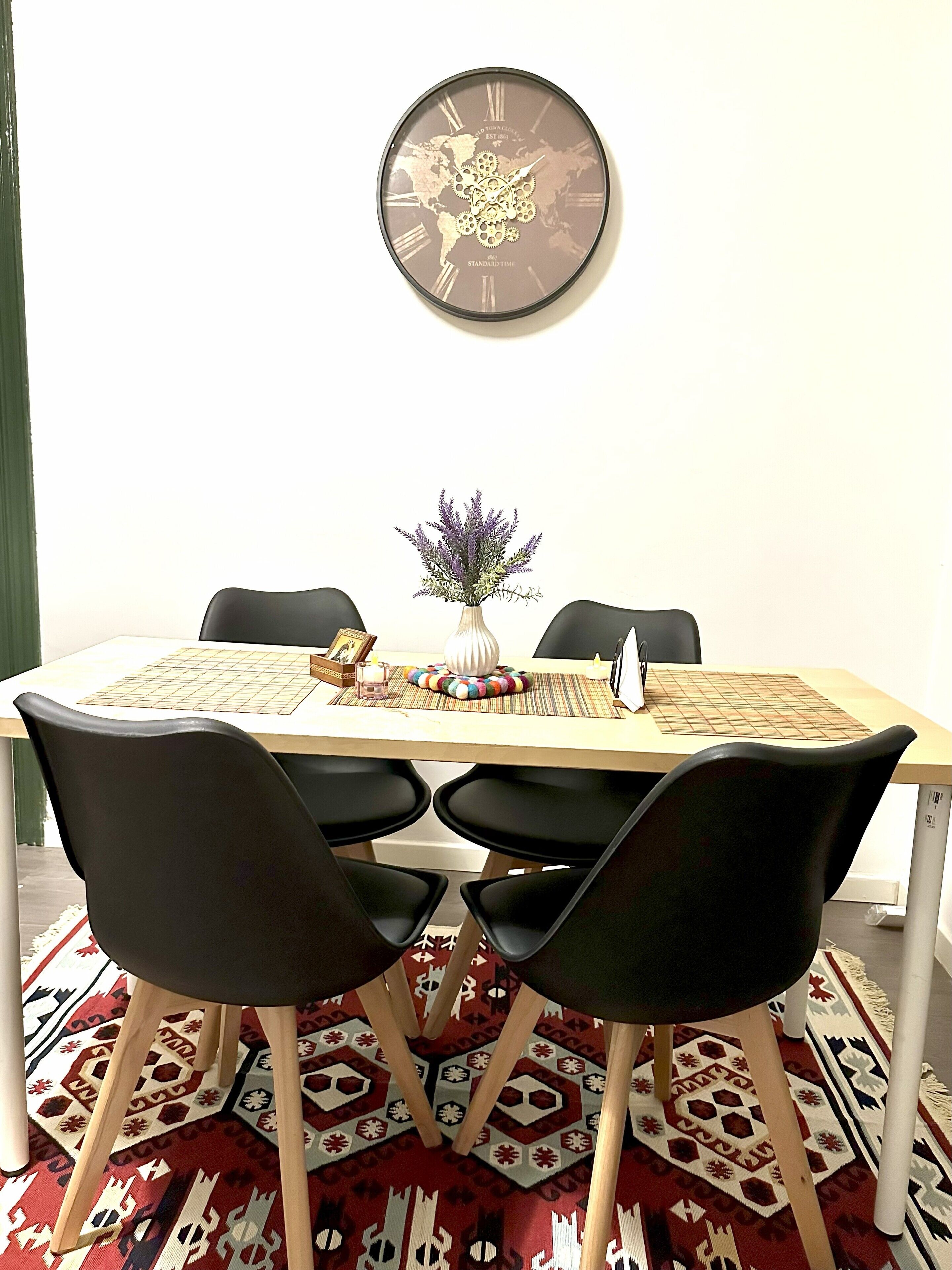 A dining area with a wooden table, black chairs, a decorative clock, and a patterned rug.