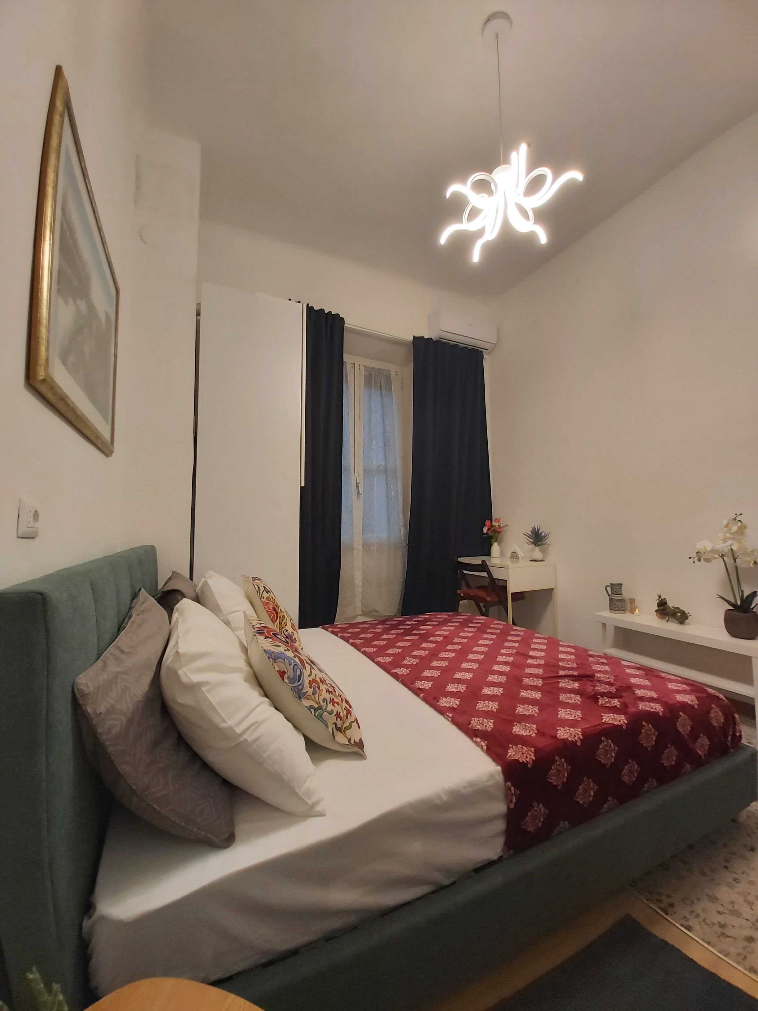 A bedroom with a bed, a bedside table, a desk, a chair, a hanging light fixture, and a window with curtains.