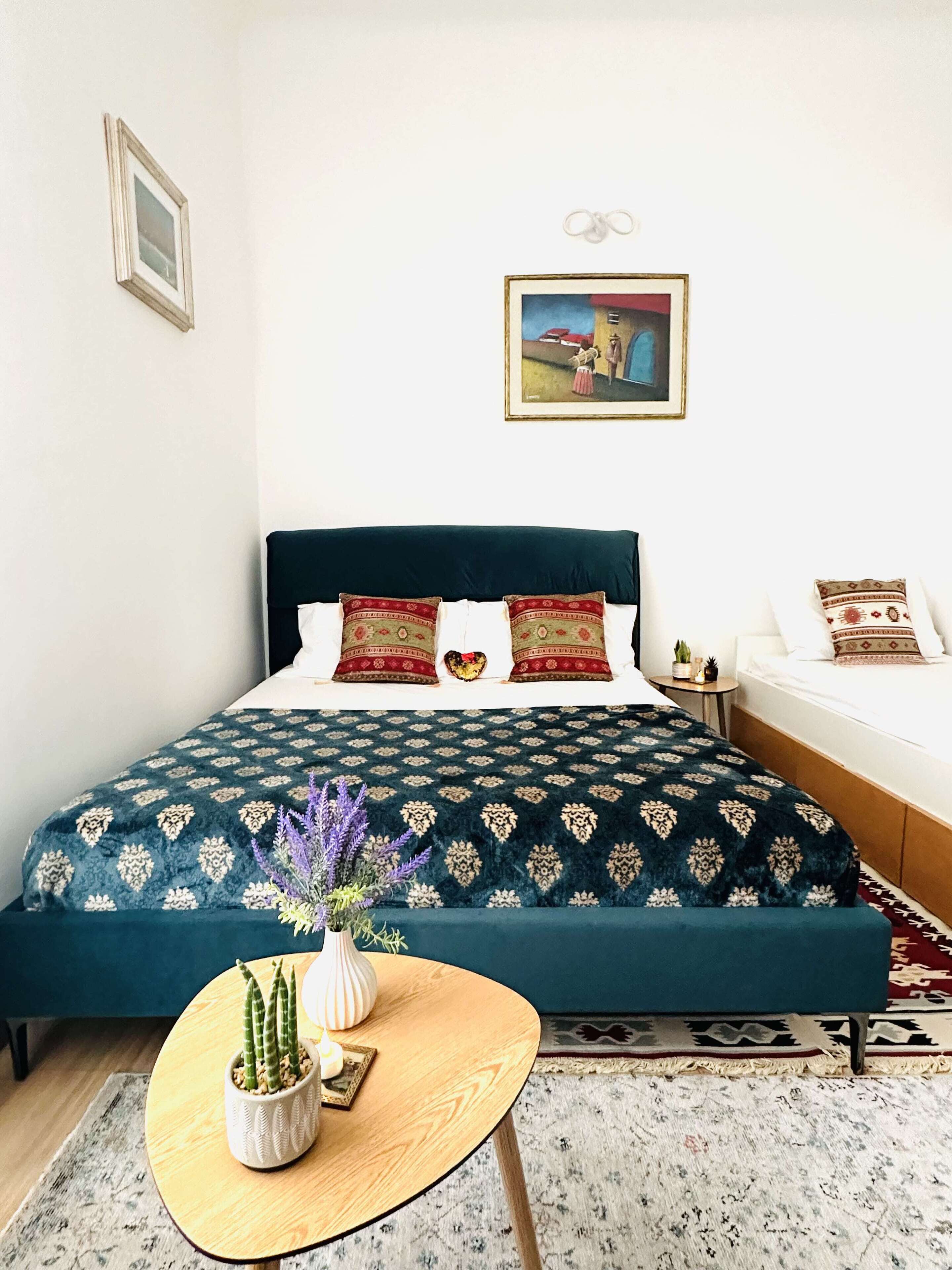 Un dormitorio con una cama grande, una mesita de noche pequeña, una mesa redonda de madera con un florero y dos cactus, y una alfombra estampada.