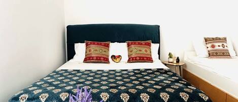 Un dormitorio con una cama grande, una mesita de noche pequeña, una mesa redonda de madera con un florero y dos cactus, y una alfombra estampada.