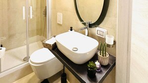 A modern bathroom with a glass-enclosed shower, a white sink with a round mirror, and a black countertop. - Caserina Lux Roma (Rome)