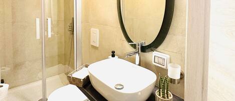 Une salle de bain moderne avec une douche à l’italienne en verre, un lavabo blanc avec un miroir rond et un plan de toilette noir.