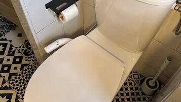 Un inodoro con asiento y tanque blancos, un dispensador de papel higiénico y un suelo con diseño.