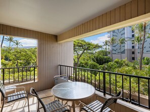En balkong med ett runt glasbord och fyra stolar, med utsikt över en frodig trädgård och en byggnad med en fasad som har ett lövmönster.