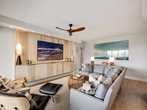 Ein modernes Wohnzimmer mit grauem Sofa, einem schwarzen Ledersitzhocker, einem Holztisch und einem Deckenventilator.