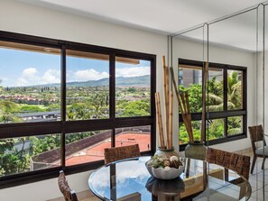A dining area with a glass table, wicker chairs, and a bowl of food. Large windows offer a view of a cityscape and greenery. - Kaanapali Alii 264 - Professionally Managed (Lahaina)