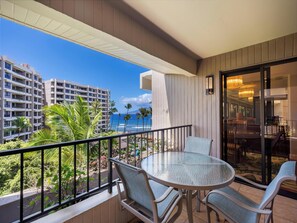 A balcony with a glass table and chairs, overlooking a beach and high-rise buildings. - 6th Floor Ocean Views - Professionally Managed (Lahaina)