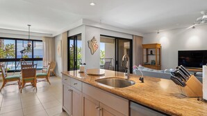 A modern kitchen with a granite countertop, stainless steel sink, and a Bosch dishwasher. There is a dining area with wicker chairs and a view of the ocean. - Wake Up With Ocean Views - Professionally Managed (Lahaina)
