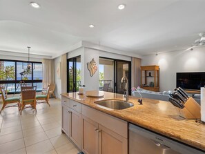 A modern kitchen with a large island, a dining area with wicker chairs, a view of the ocean, and a living room with a flat-screen TV. - Wake Up With Ocean Views - Professionally Managed (Lahaina)