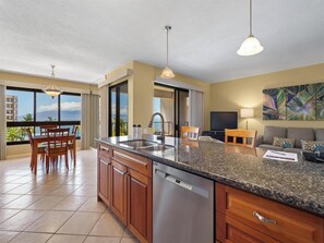 A modern kitchen with a granite countertop, stainless steel appliances, and a dining area with a view. - Wake Up With Ocean Views - Kaanapali Alii 281 (Lahaina)