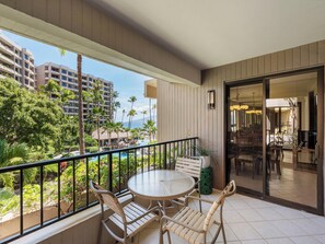 A balcony with a round table and chairs, overlooking a pool and lush greenery. - Inviting Pool & Ocean Views - Kaanapali Alii 132 (Lahaina)