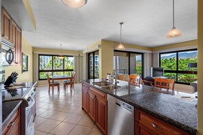 A modern kitchen with granite countertops, stainless steel appliances, and wooden cabinets. There is a dining area with a table and chairs, and large windows offering a view of greenery and buildings. - Walk to Waves, Snorkeling, Golf & Restaurants (Lahaina)