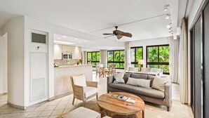 Ein geräumiger Wohnbereich mit einer Couch, einem Couchtisch und einem Esstisch. Im Hintergrund befindet sich eine Küche mit einer Mikrowelle und einem Deckenventilator.